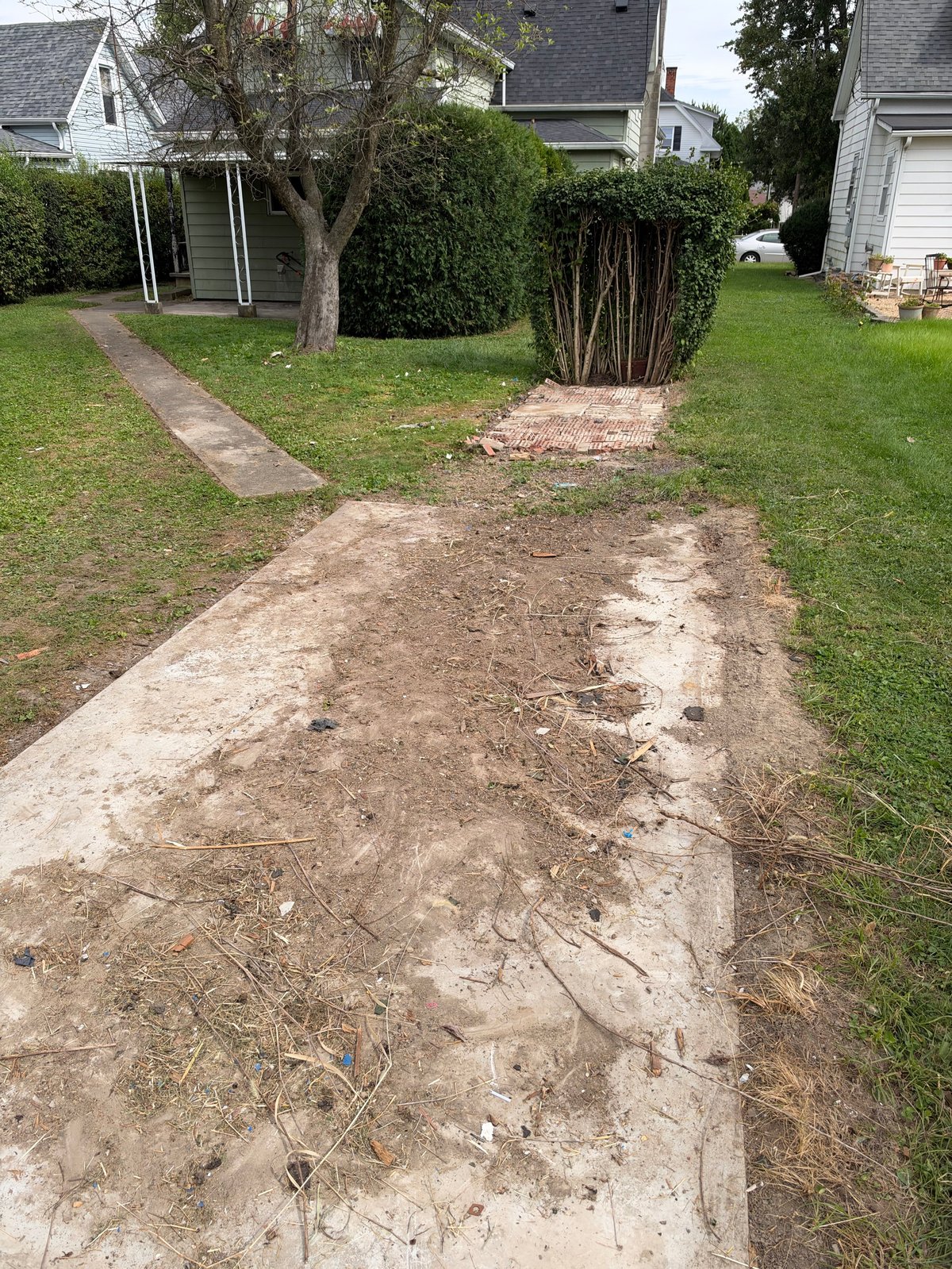 Deteriorated concrete driveway with cracked, uneven surface alongside green grass and shrubs in residential setting