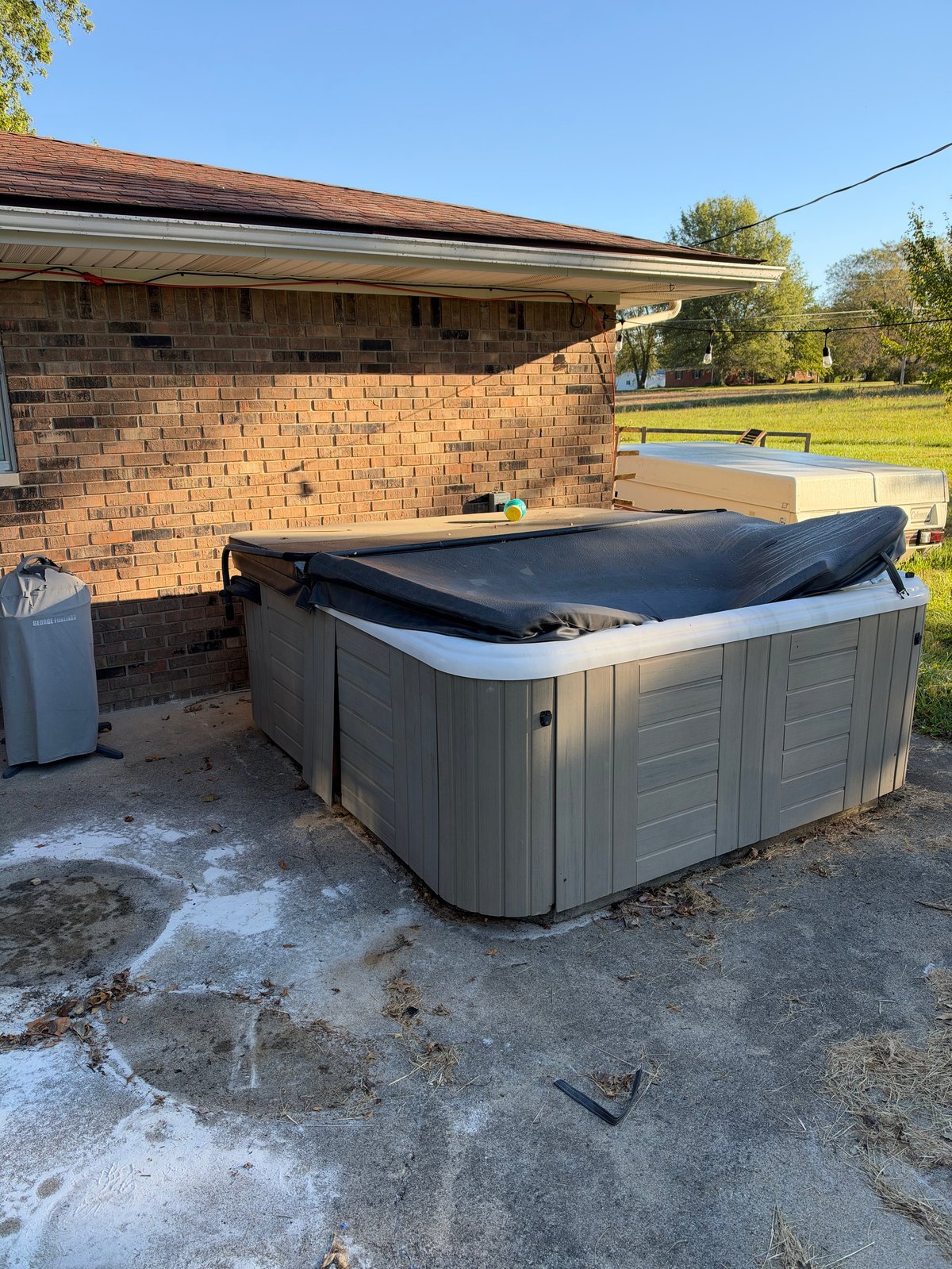 Gray hot tub with black cover outside brick building on concrete patio with green yard visible in background