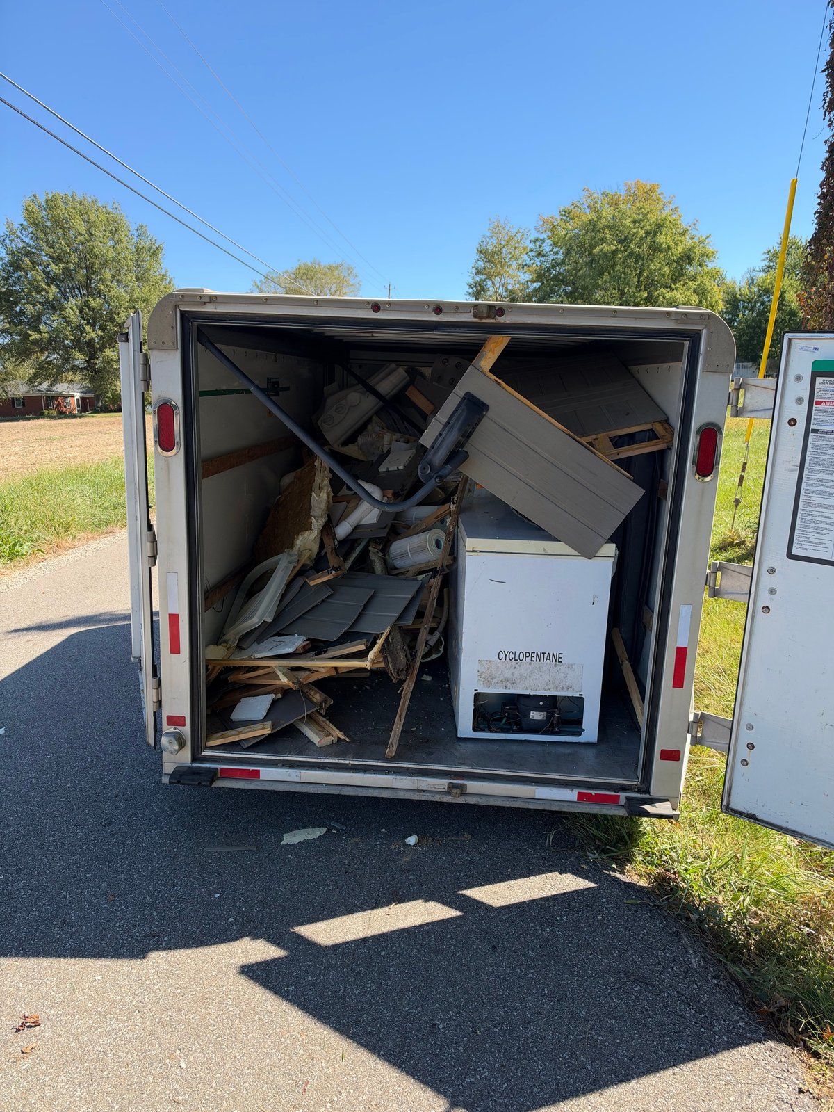 Open white cargo trailer filled with miscellaneous junk, furniture, and appliances parked outdoors on a sunny day