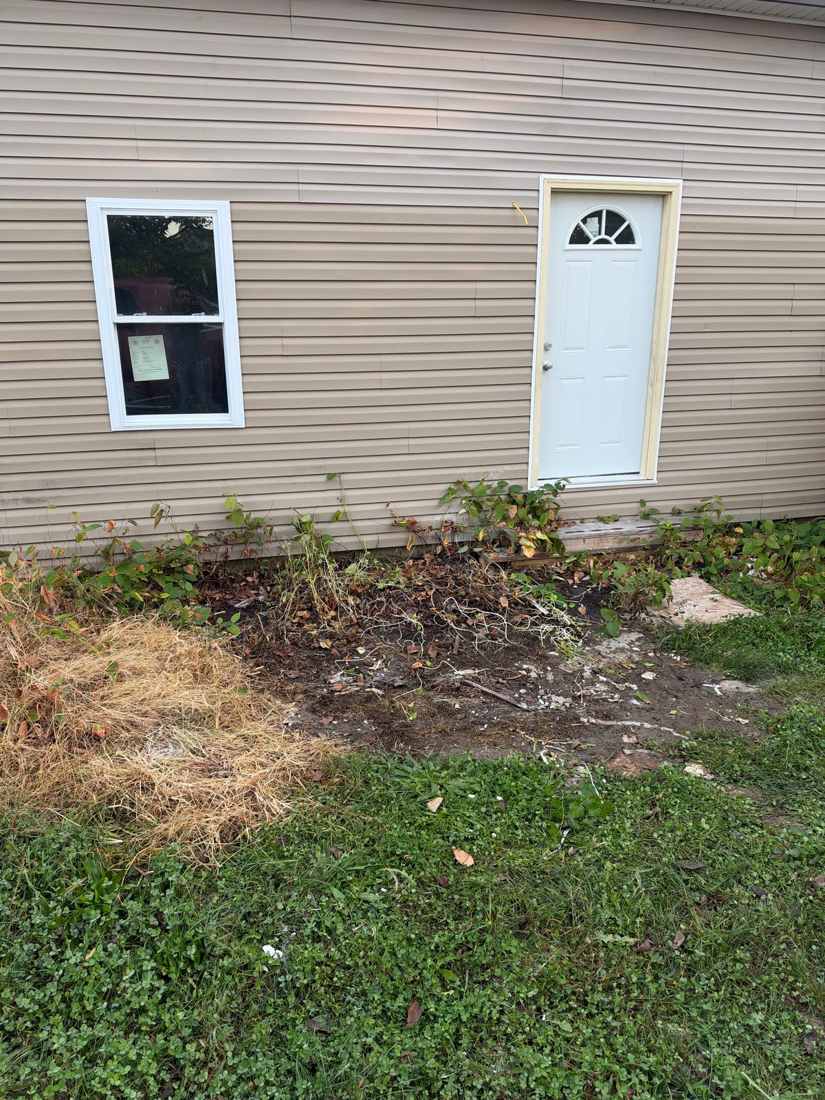 Beige metal-sided house with white window and door, featuring a cleared garden bed below after junk removal