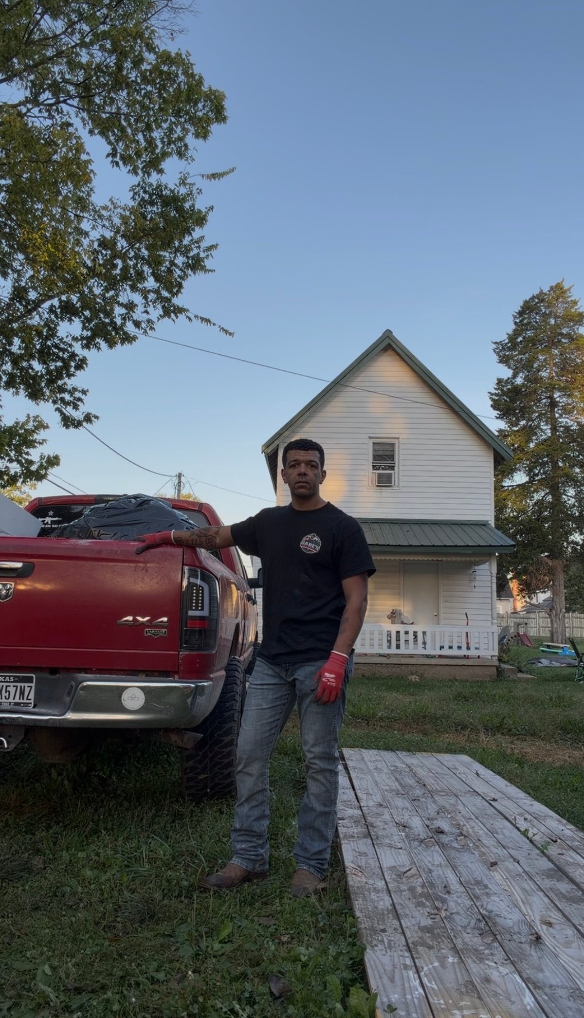 Man in black shirt standing next to red truck in front of white two-story house with green lawn