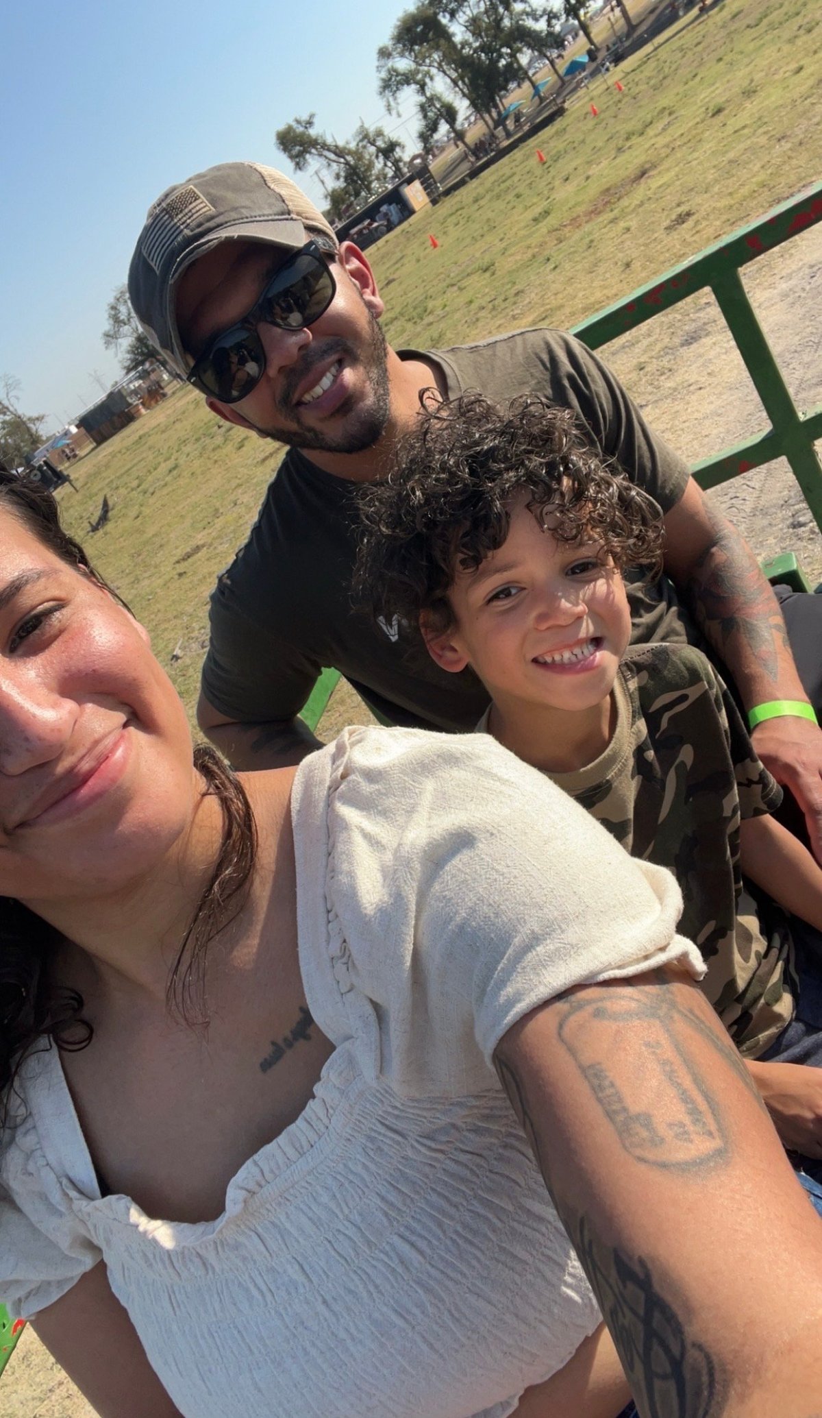 Three people smiling for a selfie at an outdoor park with green grass and trees in the background on a sunny day