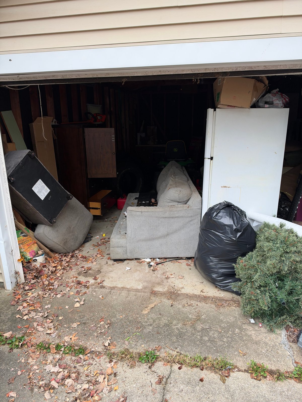 Cluttered garage interior with stored furniture, cardboard boxes, a white refrigerator, black trash bags, and assorted household items